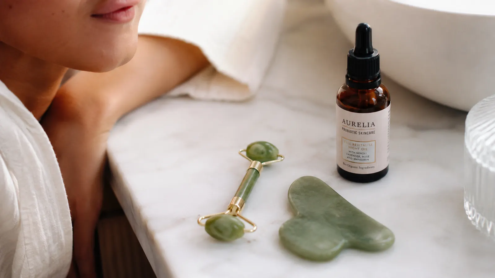 Woman in her 50s performing gua sha facial massage with a green jade tool on her cheekbone, soft morning bathroom light