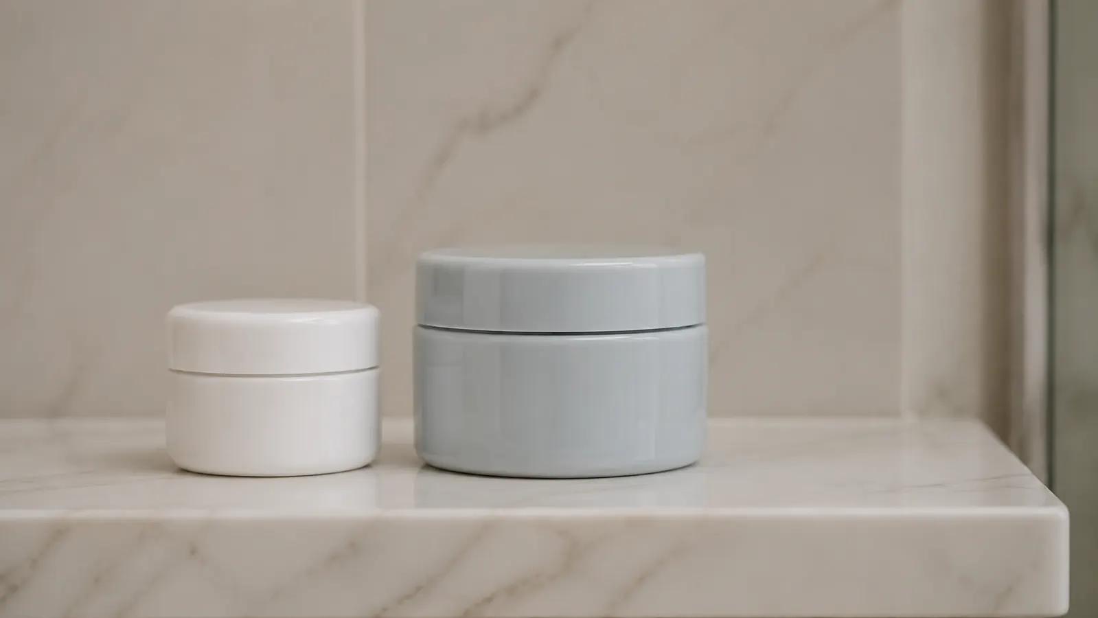 Two skincare jars of different sizes sitting side by side on a white bathroom shelf