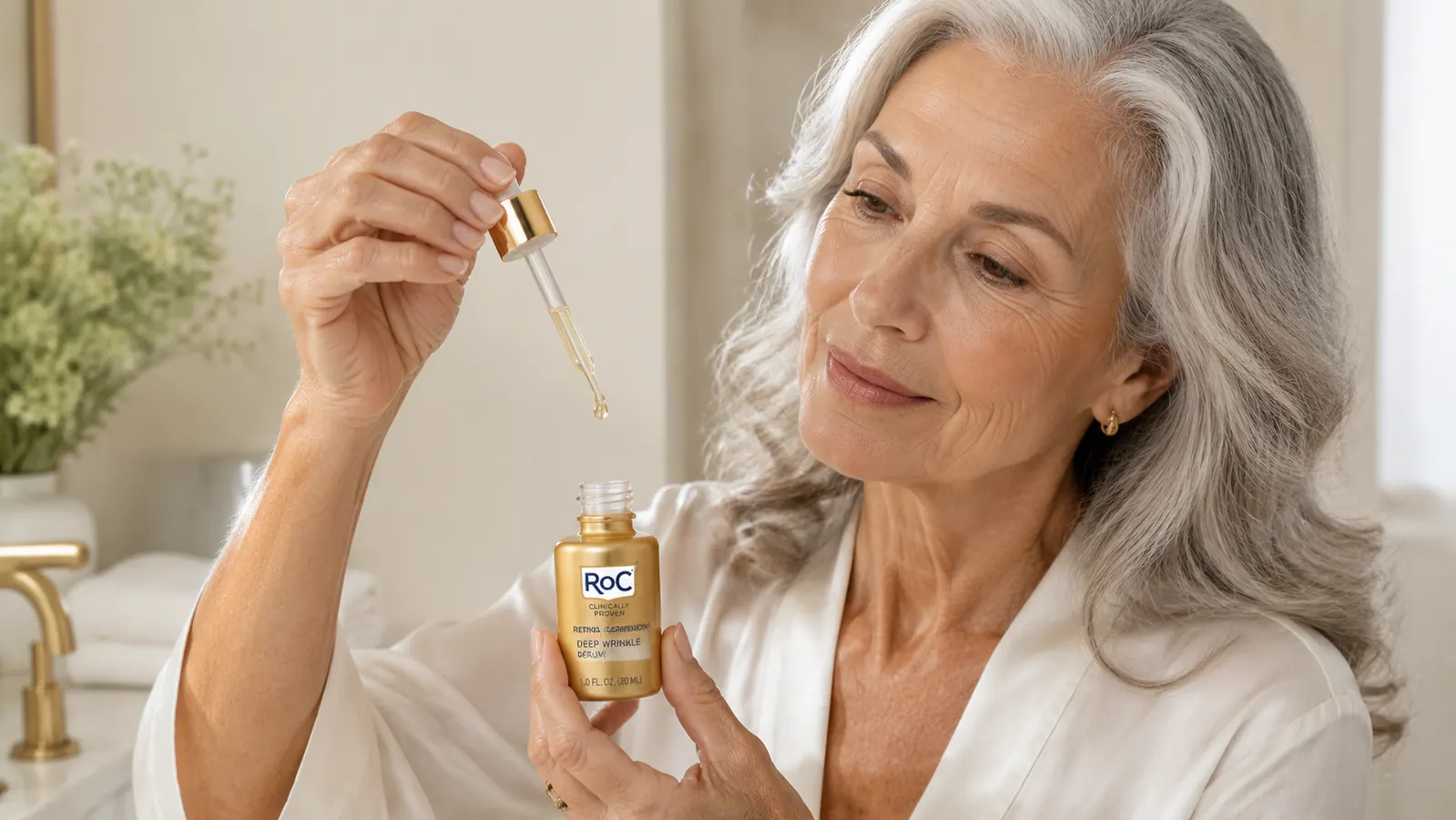 Close-up of a hand pressing a serum dropper over a small glass dish, one amber drop suspended mid-fall against a neutral background
