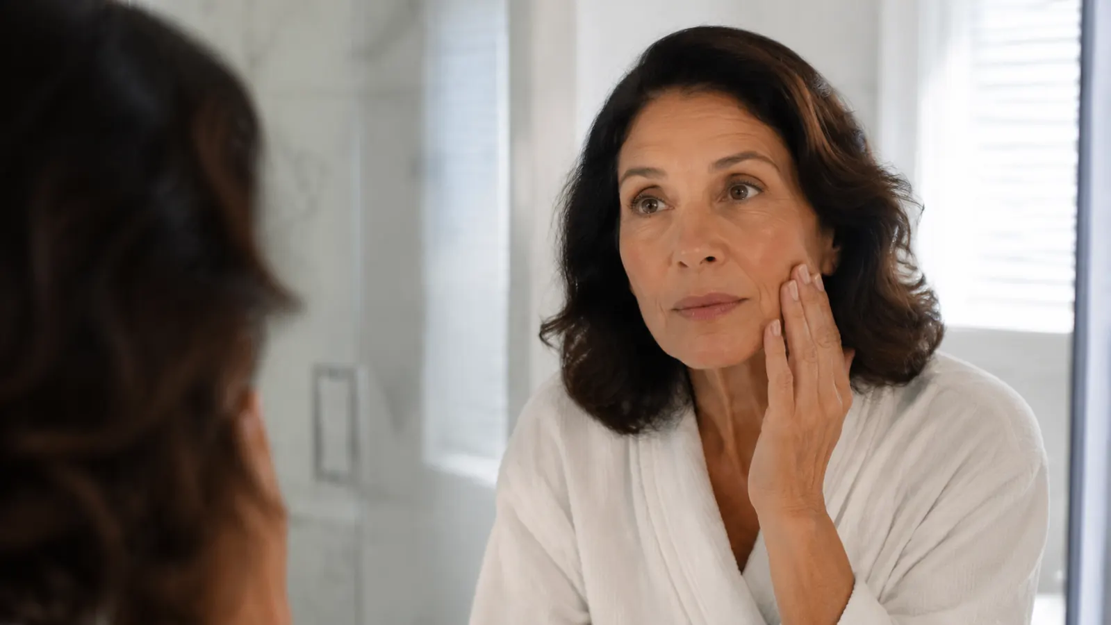 Woman in her late 50s examining her skin closely in a bathroom mirror with natural morning light
