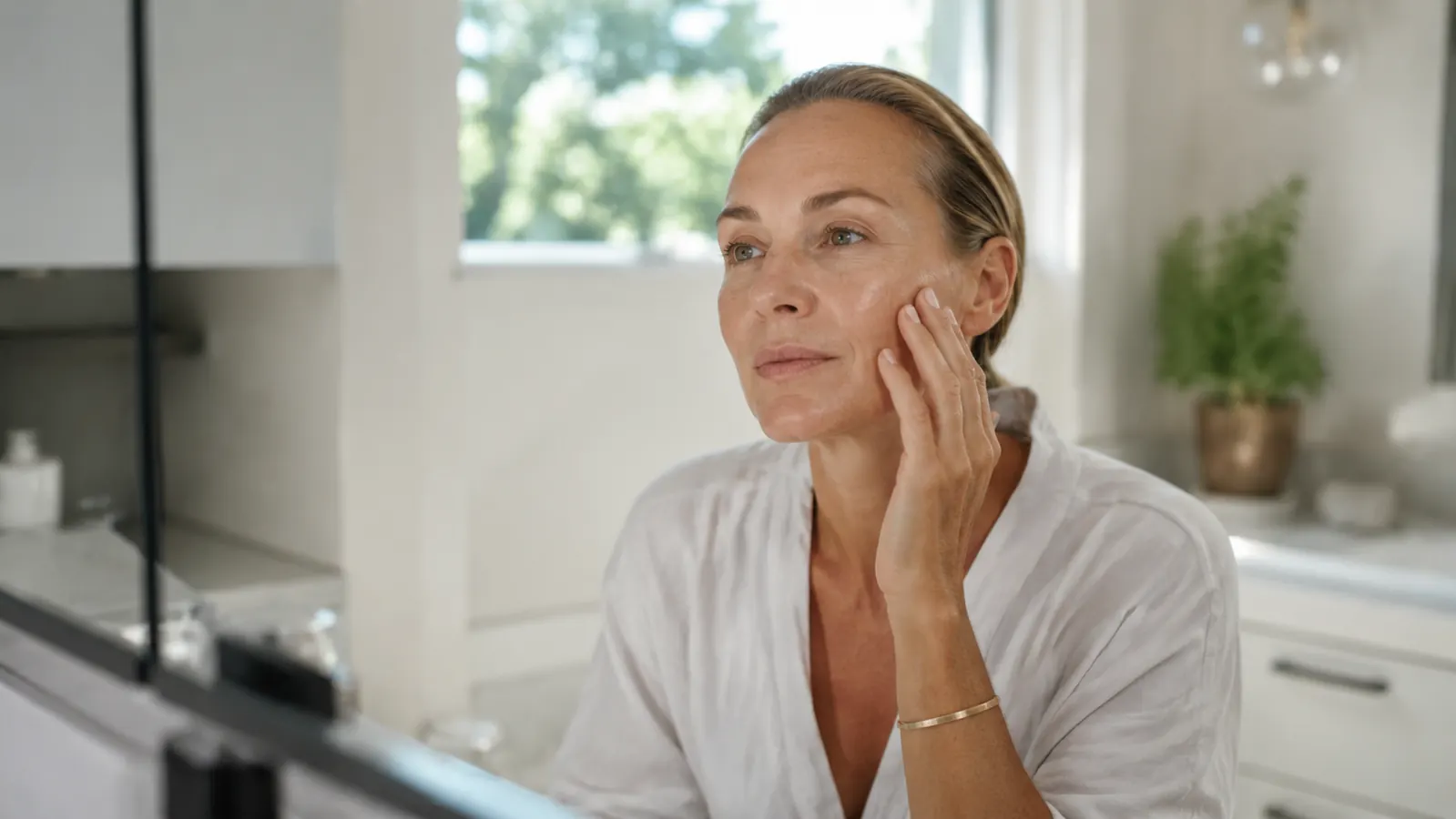 Woman in her 50s sitting at a well-lit bathroom vanity applying serum to her face in soft morning light